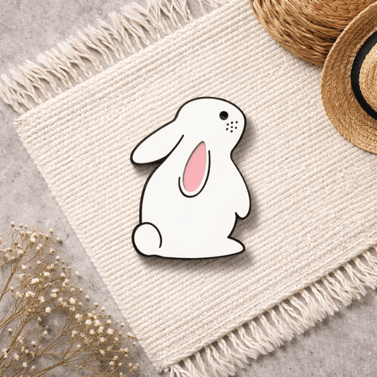 White rabbit-shaped pin on a textured fabric surface with dried flowers and a straw hat in the background.