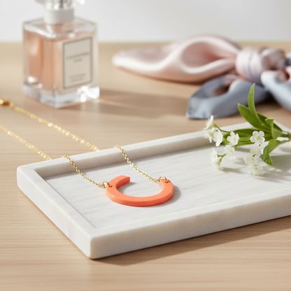 Necklace with a pink crescent moon pendant on a white tray with a bottle of perfume and flowers in the background.